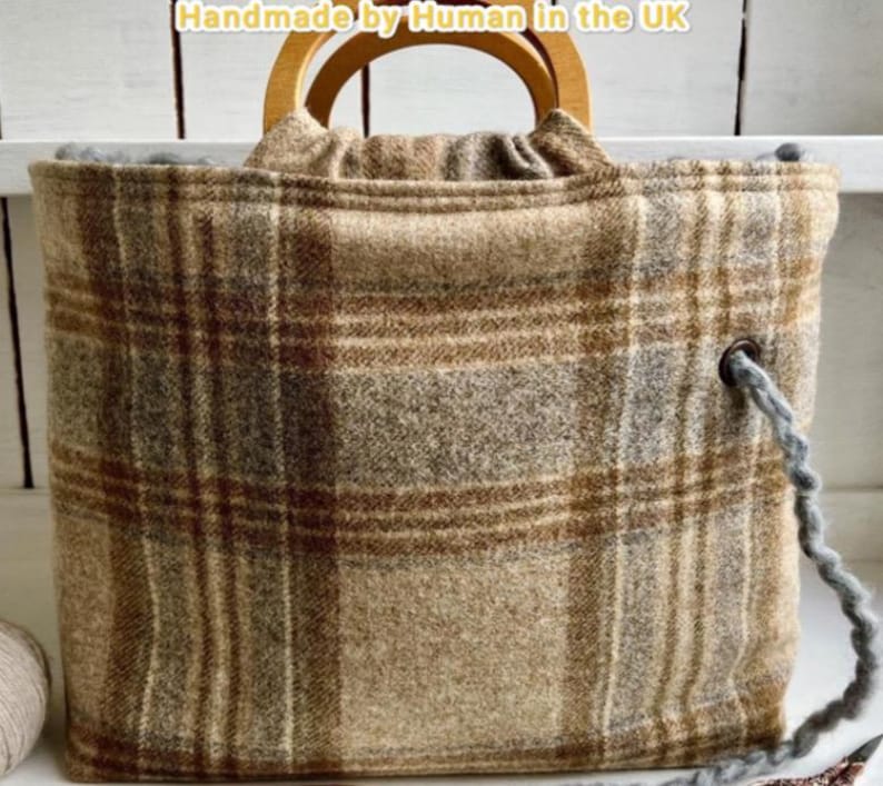 May include: A tote bag featuring a brown, tan, and gray plaid pattern with rounded wooden handles. The bag includes a gray yarn drawstring threaded through a metal eyelet on the side. The text "Handmade by Human in the UK" is visible at the top.