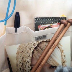 May include: A cream-colored canvas organizer bag featuring multiple pockets filled with craft supplies, including wooden knitting needles marked 9mm, a blue crochet hook, a black pen, and a small black and white polka dot notebook with a WakarAki label.