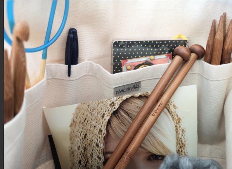 May include: A cream-colored canvas organizer bag featuring multiple pockets filled with craft supplies, including wooden knitting needles marked 9mm, a blue crochet hook, a black pen, and a small black and white polka dot notebook with a WakarAki label.