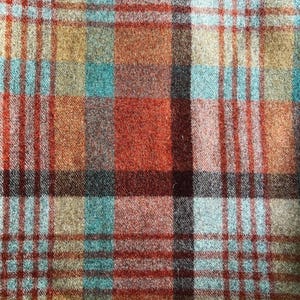 May include: A woven fabric featuring a multi-colored plaid pattern with intersecting lines in shades of burnt orange, deep red, light blue, and tan. The textile has a visible textured weave with a soft, slightly fuzzy appearance throughout.