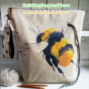 May include: A beige tote bag with a large watercolor bumblebee design. The bag has a gray drawstring, a wrist strap with a bee pattern, and the text "KnittingBagAndCase Handmade by Human in the UK". A ball of yarn and knitting needles are in front.
