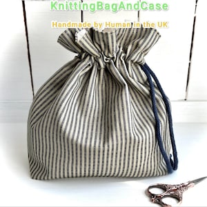 Puede incluir: Una bolsa con cordón a rayas en crema y gris oscuro, con un cordón azul marino. La bolsa es de tela y tiene el texto "KnittingBagAndCase Handmade by Human in the UK". Unas tijeras decorativas descansan cerca.