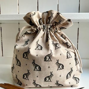 May include: A beige drawstring bag featuring a repeating pattern of black and grey hares. The bag is gathered at the top with a natural-coloured cord. Knitting needles and small scissors are visible.