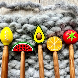 Könnte beinhalten: Fünf Stricknadeln aus Holz mit Aufsätzen in Form von Früchten: eine gelbe Zitrone, eine Wassermelonen-Scheibe, eine grüne Avocado, eine gelbe Orangenscheibe und eine rote Tomate. Die Nadeln sind vor einem grauen, strukturierten Hintergrund angeordnet.