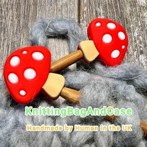 May include: Two red mushroom knitting needle stoppers with white spots and wooden stems on grey yarn. The text "KnittingBagAndCase Handmade by Human in the UK" is visible below the mushrooms.