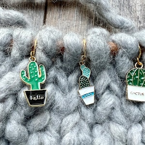 May include: Three gold cactus stitch markers with green and white enamel. The cactus on the left has the word "hello" on the pot. The middle cactus has the word "love" on the pot. The cactus on the right has the word "cactus" on the pot.