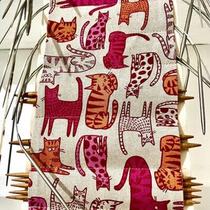 Peut inclure: Une housse à aiguilles à tricoter beige avec un motif répété de chats stylisés roses et oranges. La housse contient plusieurs aiguilles à tricoter. Le texte "Knitting Bag And Case" et "Handmade by Human in the UK" est visible.
