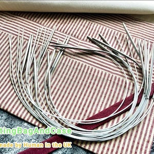 Puede incluir: Agujas de tejer circulares plateadas dispuestas sobre una tela a rayas rojas y blancas. Las agujas son curvas. La tela forma parte de un estuche o bolsa, con una cinta burdeos y una parte superior beige.
