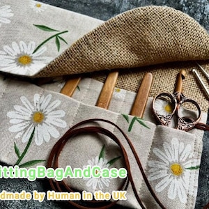 May include: A beige fabric knitting needle case with a daisy pattern, holding wooden needles, scissors, and crochet hooks. The case has a burlap flap and a brown leather cord. Text on the case reads "KnittingBagAndCase" and "Handmade by Human in the UK."