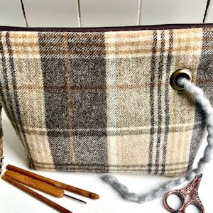 Op de afbeelding: Een geruite stoffen etui in beige en bruin met een bruine rits en bijpassende polsband. De tas heeft een messing oogje met een grijze garenlus. Haaknaalden en een schaar zijn ook zichtbaar.