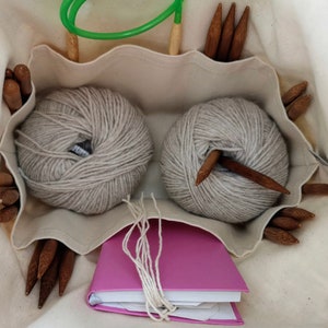 May include: A cream-colored canvas project bag containing two round balls of light gray yarn, a bright pink notebook with white yarn strands, and multiple dark brown wooden knitting needles tucked into interior pockets.