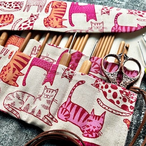 Könnte beinhalten: Eine offene Häkelnadel- und Nadelmappe mit einem Muster aus rosa und orangefarbenen Katzen auf beigem Stoff. Sie enthält Häkelnadeln, Stricknadeln und eine dekorative Schere. Zwei Häkelnadeln mit grünen und pinkfarbenen Griffen sind ebenfalls zu sehen. Für Handarbeiten.