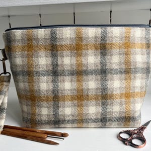 Op de afbeelding: Een rechthoekige stoffen tas met een crème, grijs en goud geruit patroon en een polsbandje. Naast de tas liggen houten breinaalden, een haaknaald en een decoratieve schaar.