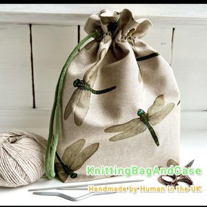 May include: Beige drawstring bag with a dragonfly print, green drawstring, and the text "KnittingBagAndCase Handmade by Human in the UK". A ball of yarn and knitting needles are next to the bag.