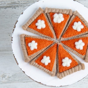 May include: A white plate with seven felt pumpkin pie slices. Each slice has a brown crust and an orange filling with a white felt dollop of whipped cream.