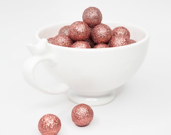 May include: A white teacup filled with rose gold glitter balls. The teacup is on a white surface. Two rose gold glitter balls are in front of the teacup. The text "Rose Gold" is written below the balls.