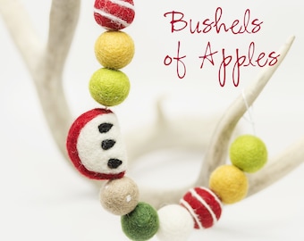 May include: A garland of felt balls in red, white, green, and yellow with a felt apple slice. The garland is hanging from a white antler and the text "Bushels of Apples" is written in red.