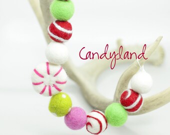 May include: A garland of felt balls in red, white, green, and pink, resembling candy canes and peppermint candies. The garland is strung on a white cord and is hanging from a white antler.