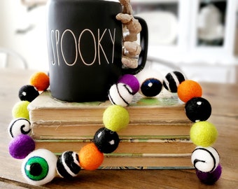 May include: A black ceramic mug with the word "SPOOKY" written on it, a skeleton hand as the handle, and a colourful felt ball garland with black and white stripes, green eyes, orange, purple, and lime green balls. The garland is draped over a stack of books.