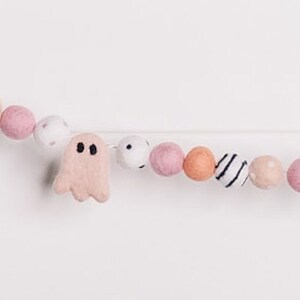 May include: A pink and white felt garland with ghosts and bats. The garland is strung on a white string and has pink, white, and orange felt balls.