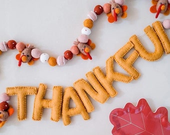 May include: A felt garland with a string of orange, pink, and white pom poms and felt turkeys. The garland is hanging above a felt banner that says "THANKFUL" in yellow letters.
