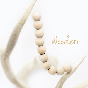Pu&ograve; includere: Una collana di perline di legno naturale &egrave; appesa a un palco di cervo bianco. Le perline sono rotonde e lisce, e la corda &egrave; legata al palco con un semplice nodo. Il testo "Wooden" &egrave; scritto in lettere dorate sotto le perline.