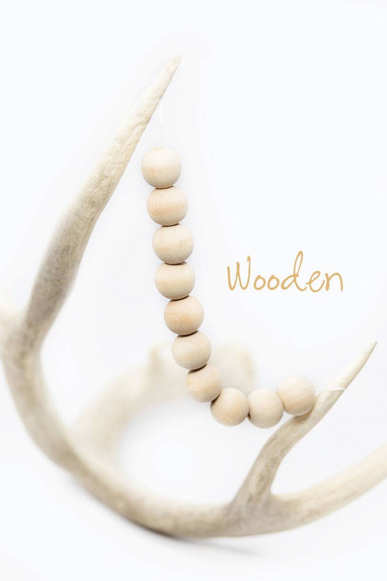 Pu&ograve; includere: Una collana di perline di legno naturale &egrave; appesa a un palco di cervo bianco. Le perline sono rotonde e lisce, e la corda &egrave; legata al palco con un semplice nodo. Il testo "Wooden" &egrave; scritto in lettere dorate sotto le perline.
