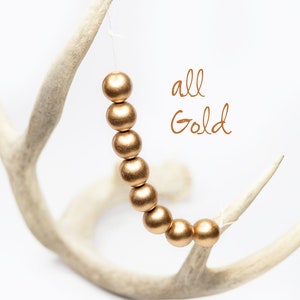 Pu&ograve; includere: Una collana di perle dorate pende da un corno di cervo bianco. Le perle sono rotonde e hanno una finitura opaca. Il testo "all Gold" &egrave; scritto in corsivo sotto le perle.