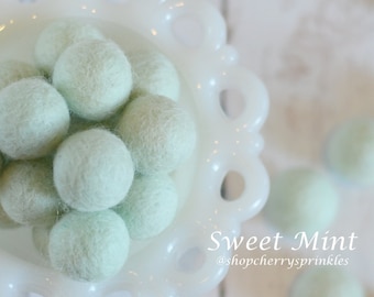 May include: A white bowl filled with soft, light green felt balls. The bowl has a decorative, scalloped edge. The text "Sweet Mint @shopcherryssprinkles" is visible below the bowl.