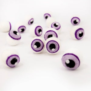 May include: A group of white felt eyeballs with purple eyelids and black pupils. The eyeballs are scattered on a white surface.