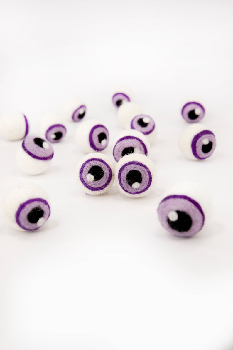 May include: A group of white felt eyeballs with purple eyelids and black pupils. The eyeballs are scattered on a white surface.