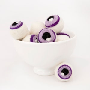 May include: A white bowl filled with purple and white felt eyeballs. The eyeballs have black pupils and are arranged in a pile.