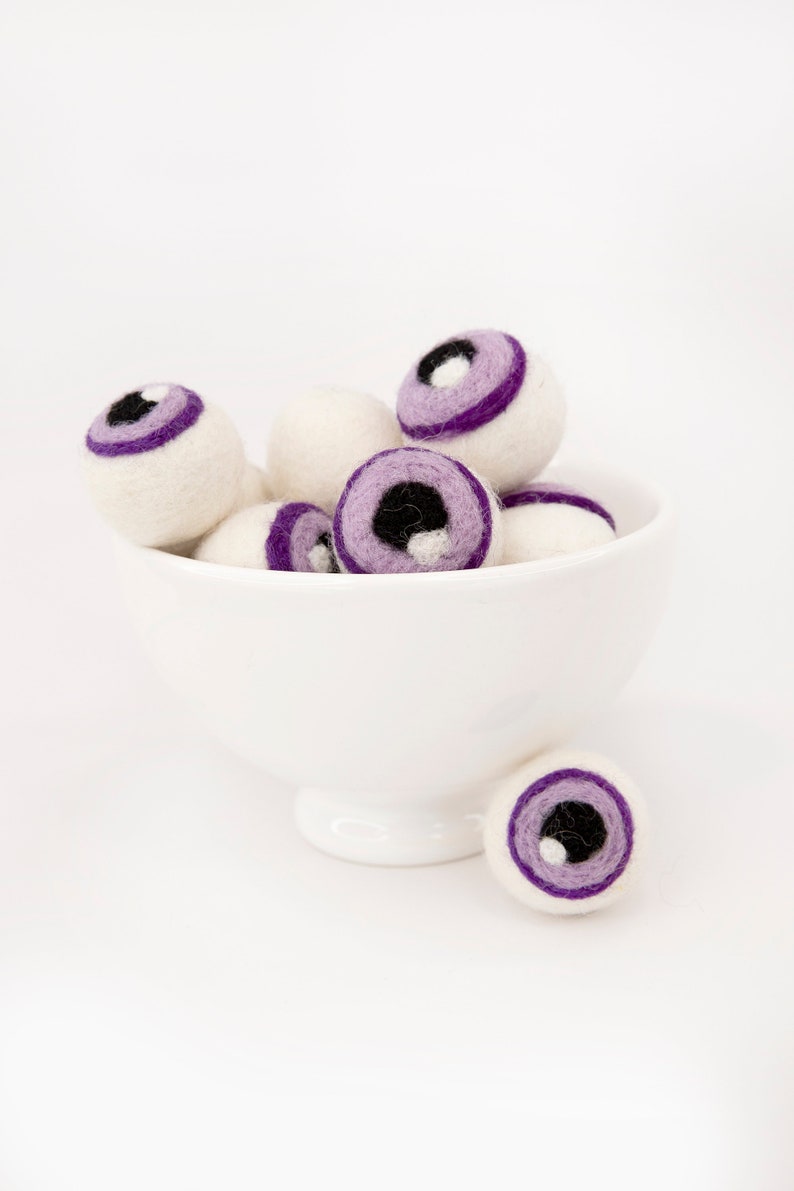 May include: A white bowl filled with purple and white felt eyeballs. The eyeballs have black pupils and are arranged in a pile.