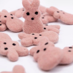 May include: A group of pink felt bunny shapes with black eyes. The bunnies are arranged in a cluster on a white background.