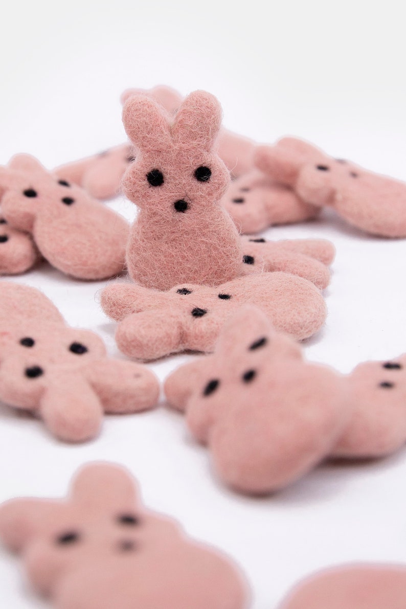 May include: A group of pink felt bunny shapes with black eyes. The bunnies are arranged in a cluster on a white background.
