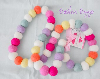 May include: A garland of colorful felt balls in shades of pink, purple, blue, yellow, orange, and white. The garland is arranged in a circle and says "Easter Eggs" in purple text.