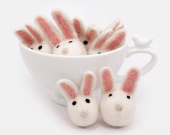May include: A white ceramic teacup filled with small white felt bunny rabbit figurines with pink ears. The bunnies are arranged in a cluster, with their heads peeking out of the cup.