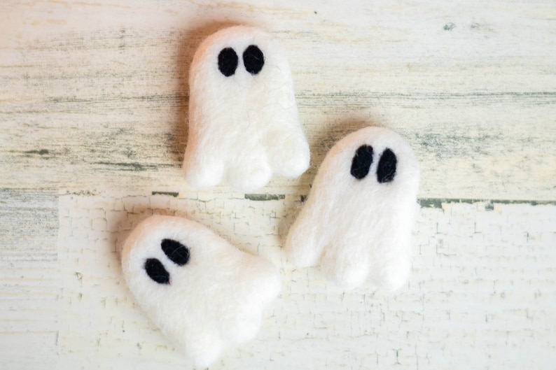 May include: Three white felt ghost ornaments with black felt eyes. The ghosts are arranged on a white wood background.