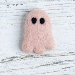 May include: A small, pink felt ghost with black eyes. The ghost is sitting on a white wooden surface.