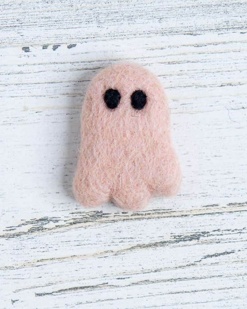 May include: A small, pink felt ghost with black eyes. The ghost is sitting on a white wooden surface.