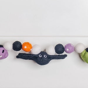Pu&ograve; includere: Una ghirlanda di Halloween con palline di feltro in bianco, nero, arancione, verde e viola. La ghirlanda presenta ritagli di feltro di un cappello da strega, un fantasma, un ragno, un Frankenstein e un occhio.