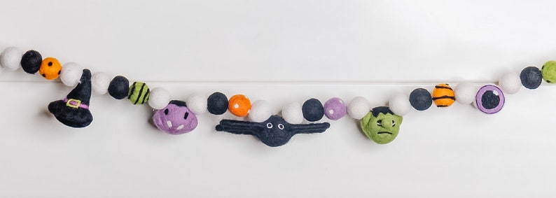 Pu&ograve; includere: Una ghirlanda di Halloween con palline di feltro in bianco, nero, arancione, verde e viola. La ghirlanda presenta ritagli di feltro di un cappello da strega, un fantasma, un ragno, un Frankenstein e un occhio.