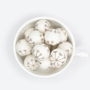 Puede incluir: Un taz&oacute;n de cer&aacute;mica blanco lleno de bolas de fieltro blanco, cada una con un dise&ntilde;o de diente de le&oacute;n marr&oacute;n.