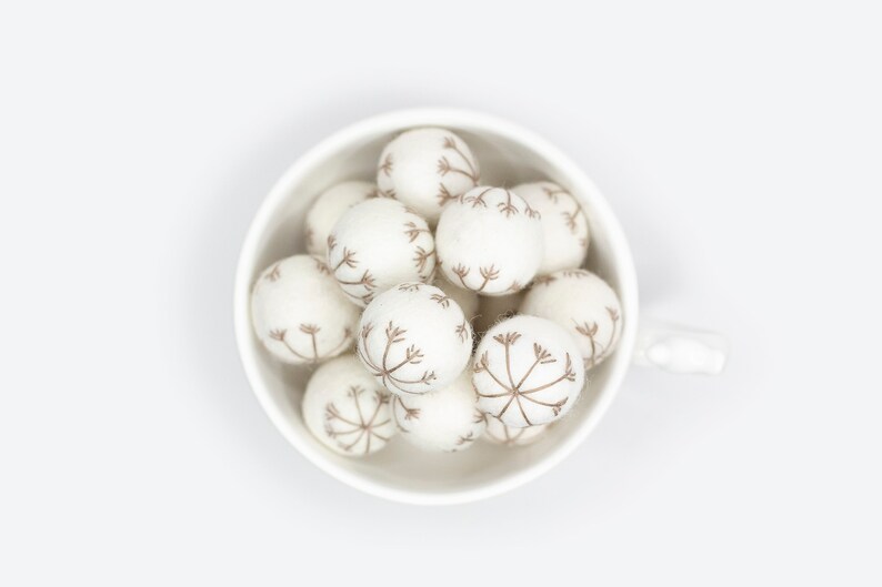 Puede incluir: Un taz&oacute;n de cer&aacute;mica blanco lleno de bolas de fieltro blanco, cada una con un dise&ntilde;o de diente de le&oacute;n marr&oacute;n.