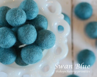 May include: A white bowl filled with teal blue felt balls. The bowl has a decorative edge. The text "Swan Blue @shopcherry sprinkles" is below the bowl.