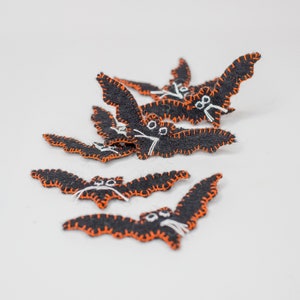 May include: A collection of black felt bat decorations with orange stitching and white facial details. These Halloween-themed bats are arranged on a white surface, showcasing their handmade quality and festive design.