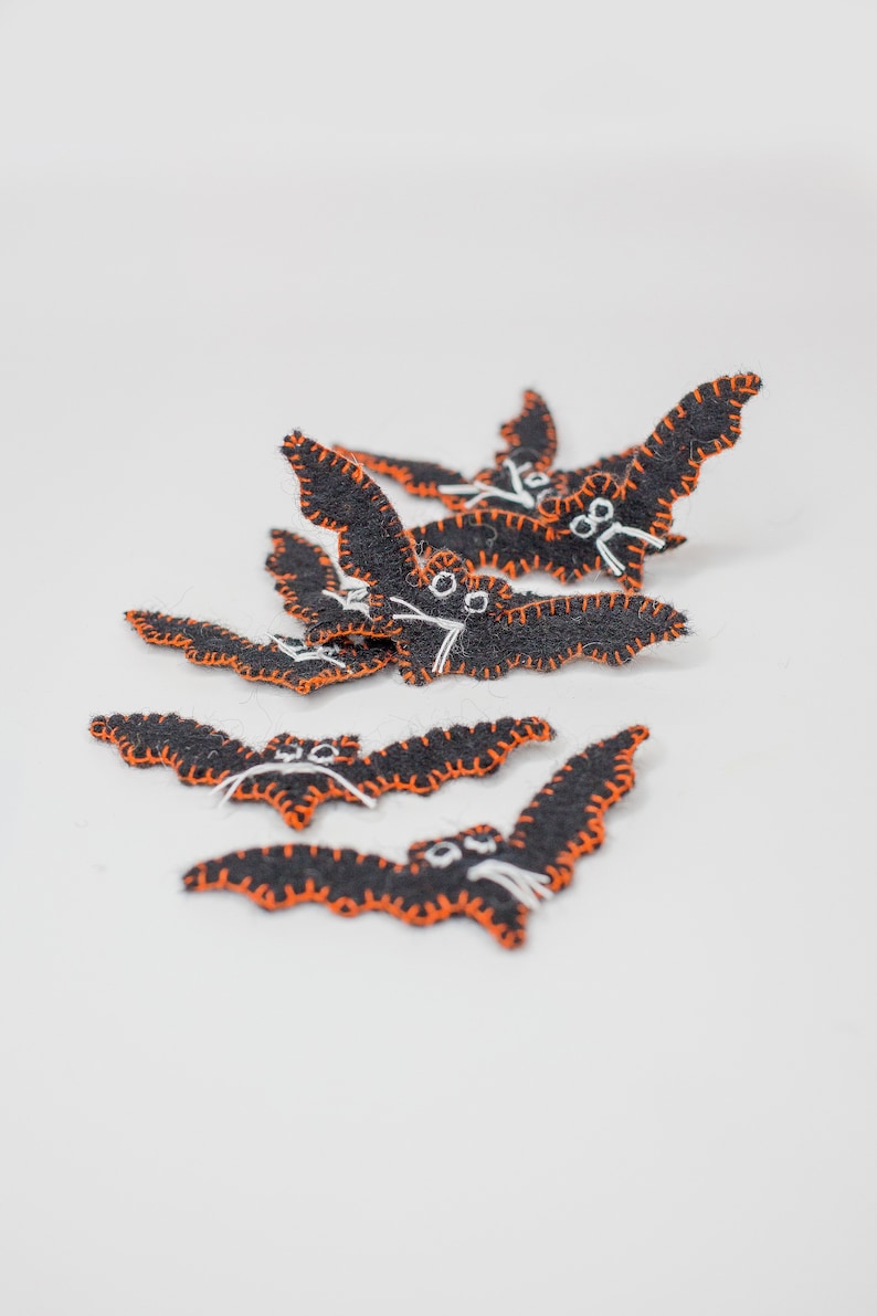 May include: A collection of black felt bat decorations with orange stitching and white facial details. These Halloween-themed bats are arranged on a white surface, showcasing their handmade quality and festive design.