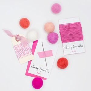 May include: A flat lay of pink and red felt balls, pink and white twine, and two cards that say "Shop Cherry Sprinkles" with an Etsy logo.