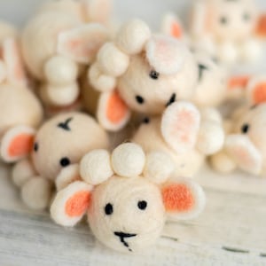 May include: A group of small, white, felt sheep with pink ears and black eyes. The sheep are arranged in a cluster on a white wooden surface.