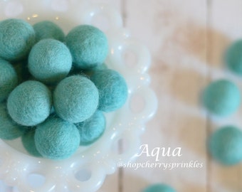May include: A bowl filled with aqua blue felt balls. The bowl is white with a scalloped edge. The felt balls are soft and fluffy. The text "Aqua" and "@shopcherry sprinkles" is written below the bowl.